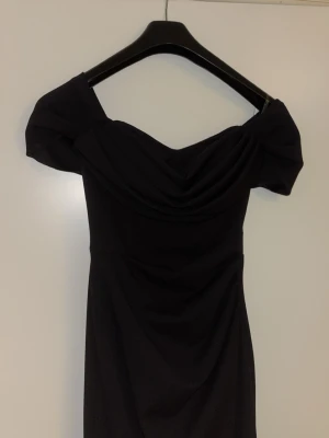 Svart offshoulder klänning - Säljer en elegant svart offshoulder klänning med draperad urringning och figurnära passform. Klänningen är kort och har en stilren design som framhäver axlarna. Perfekt för dig som vill ha en klassisk och trendig look.