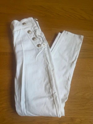 Vita högmidjade byxor med knappar - Vita högmidjade byxor från Guess i jeansmaterial med dekorativa guldknappar framtill och dragkedja i sidan. Aldrig använda och med prislapp kvar, stl 28 (se bild)