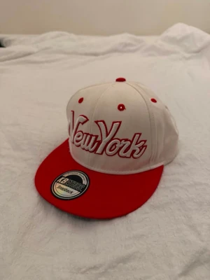 Limiterad KBETHOS vit, röd keps - Keps med texten "New York" broderad i rött på framsidan. Kepsen har en röd skärm och en vit krona med röda detaljer. Baktill finns en justerbar röd snapback-stängning. Insidan av kepsen har ett vitt svettband och märkets logotyp tryckt längs sömmarna.