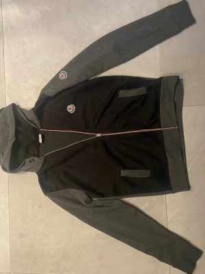Moncler hoodie i svart och grå - Snygg hoodie från Moncler i svart och grått med dragkedja framtill. Tröjan har huva, två fickor och Moncler-logga på bröstet och ärmen. Materialet är en mix av mjuk bomull och mesh-tyg på framsidan för extra stilpoäng. Märket på dragkedjan är av men dragkedjan går fortfarande att dra upp lätt och smidigt.