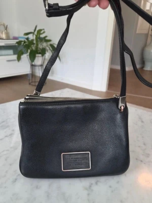 Svart axelväska Marc by Marc Jacobs - Snygg svart axelväska från Marc by Marc Jacobs i mjukt, strukturerat läder. Väskan har justerbar axelrem, silverfärgade metalldetaljer och en metallbricka med logotyp på framsidan. Perfekt storlek för det viktigaste och enkel att matcha till olika outfits.Den här svarta Marc by Marc Jacobs-väskan i läder har en tydlig rektangulär logotypplatta och dubbla dragkedjor. Fint skick, en riktig klassiker. Lite lite slitage på spännen, syns på bilden.   Mått: 24 cm bred, 16 cm hög och ca 3 cm djup.  