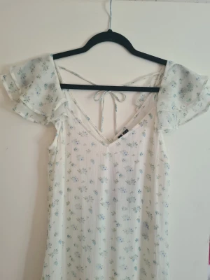 Blommig vit klänning Vero Moda - Söt vit klänning från Vero Moda med små blå blommor, volangärmar och v-ringning. Prislappen är kvar och klänningen är helt oanvänd. Klänningen har knyt i ryggen och är i ett lätt material perfekt för sommaren.  Storlek på klänningen är XS. 