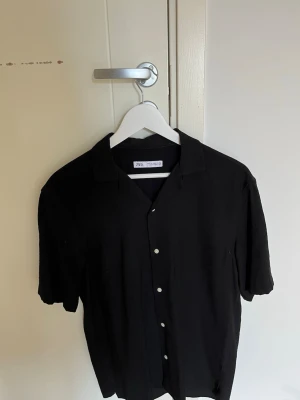 Svart kortärmad skjorta från Zara - Stilren svart skjorta från Zara med korta ärmar och klassisk krage. Skjortan har knappar framtill och är tillverkad i ett mjukt material som känns skönt mot huden. Perfekt för en avslappnad och trendig look. Aldrig använd.