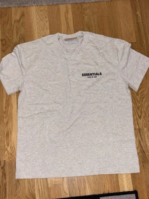Grå Essentials Fear of God t-shirt - Snygg ljusgrå t-shirt från Fear of God Essentials med svart logotyptryck både fram och bak. Klassisk rund hals och korta ärmar. Tillverkad i mjuk bomull för en skön och avslappnad passform. Nypris runt 900kr. Men säker eftersom storleken (XS) vart lite stor. Säljer därför för 650kr
