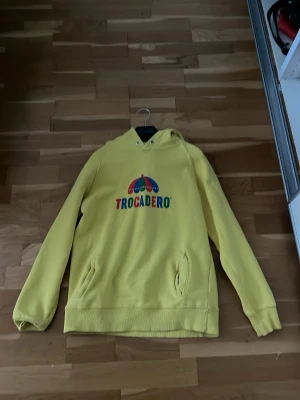 Gul Trocadero hoodie med tryck - Gul hoodie från Trocadero med färgglatt paraply-tryck och logga på bröstet. Hoodien har huva med dragsko och fickor på sidorna. Perfekt för dig som gillar unika och färgstarka plagg. Mjuk bomullskänsla och avslappnad passform.