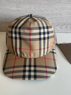 Beige rutig keps från Burberry - Snygg keps från Burberry i klassiskt beige rutigt mönster med svarta, vita och röda linjer. Kepsen har böjd skärm och justerbar rem bak. Tillverkad i bomull för en bekväm och stilren look. Perfekt accessoar för dig som gillar streetstyle och premiumkänsla.