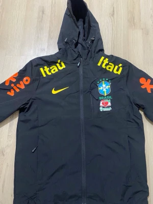 Svart Brasil Nike vindjacka - Svart vindjacka från Nike med Brasilien fotbollslandslagets emblem på bröstet. Jackan har huva, dragkedja, gula och orange loggor och sponsortryck som Itaú och Vivo. Tillverkad i lätt syntetmaterial med meshfoder för extra komfort.
