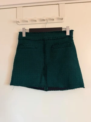 Grön kortkjol i tweed-look - Mörkgrön kortkjol med råa kanter och två dekorativa fickor framtill. Kjolen har en strukturerad tweed-liknande yta och rak passform som ger en trendig vibe. Perfekt att styla med stickad tröja eller boots. Aldrig använd. Köpt på Sellpy