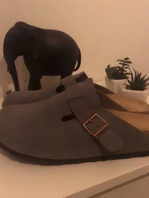Gråa Birkenstock Boston tofflor - Säljer ett par gråa Birkenstock Boston tofflor i mocka med justerbart spänne i metall. Klassisk modell med bekväm fotbädd och öppen häl. Perfekta för en chill och stilren vardagslook.