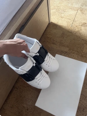 Valentino Garavani vita sneakers - Säljer ett par vita sneakers från Valentino Garavani med en bred svart rem över mitten. Skorna har klassisk låg profil, vita snören och detaljer i guld på plösen. Tillverkade i slätt läder för en clean och lyxig look.
