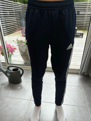 Mörkblå Adidas träningsbyxor - Modellen är 167. Säljer ett par mörkblå träningsbyxor från Adidas med vita detaljer och logga på låret. Byxorna har smal passform, resår i midjan och klassiska ränder längs sidorna. De har en gradkedja där nere på sidan av benet. Perfekta för dig som gillar sportig stil.