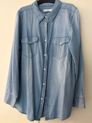 Blå jeansskjorta från XLNT - Snygg blå jeansskjorta från XLNT, KappAhl,  med klassisk krage, långa ärmar och två bröstfickor med knappar. Skjortan har en avslappnad passform och är tillverkad i mjukt bomullsmaterial. Perfekt att styla lager på lager. Storlek L i XLNT = 44/46.
