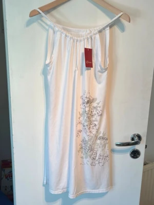 Vit blommig klänning från Guess - Säljer en vit axelbandslös klänning från Guess med blommigt mönster i grått och grönt på framsidan. Klänningen har en avslappnad passform och är gjord i mjuk bomull, perfekt för varma dagar. Snygg och enkel design med diskret logga.