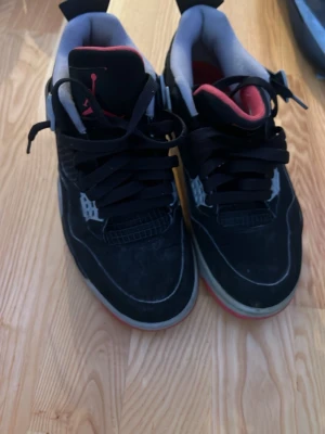 Svarta Air Jordan 4 Retro sneakers - Säljer ett par svarta Air Jordan 4 Retro sneakers med röda och grå detaljer. Skorna har meshpaneler, plastdetaljer och tjock sula. Insidan är ljusgrå och plösen har en röd Jumpman-logga. Klassisk och ikonisk modell för dig som gillar streetwear.