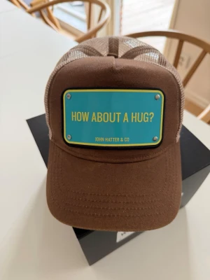 John Hatter & Co – “How About A Hug?” Truckerkeps - Säljer en unik och iögonfallande keps från John Hatter & Co med det charmiga budskapet “How About A Hug?”. Perfekt för dig som gillar att sticka ut med stil och samtidigt sprida lite positiv energi.  Original från John Hatter & Co Trucker-modell med ventilerande mesh baktill.  Justerbar storlek för optimal passform Snygg brun/beige färgkombination Mycket fint skick  