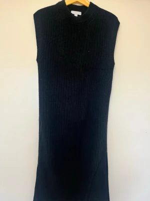 Svart ribbad stickad långklänning - Säljer en svart stickad långklänning  med ribbad struktur och hög hals. Klänningen är ärmlös och har en rak, enkel passform som ger en clean look. Perfekt att styla med boots eller sneakers för en trendig vibe. Där är två slitsar en på var sida dock har jag sytt igen de men sömmarna går agg få upp om man vill. Använd 2 gånger 