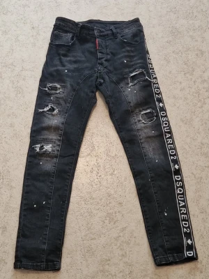 Svarta Dsquared2 jeans med tryck - Säljer ett par svarta Dsquared2 jeans med slitningar, hål och vita färgstänk. Snygg detalj med Dsquared2-tryck längs ena benet. Jeansen har smal passform och är perfekta för dig som vill sticka ut med en edgy streetstyle.