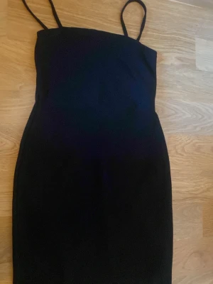 Svart tight klänning med tunna band - En enkel och stilren svart klänning med smala axelband och figurnära passform. Klänningen är kort och har en minimalistisk design som passar perfekt till många olika tillfällen. Materialet känns mjukt och stretchigt.