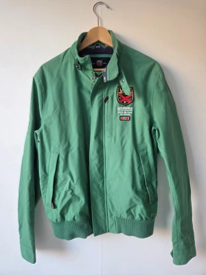 Grön vindjacka från Morris - Snygg grön vindjacka från Morris med broderad patch och detaljer på bröstet. Jackan har hög krage med spänne, dragkedja och tryckknappar samt ribbade muddar. Perfekt för en sportig och stilren look.
