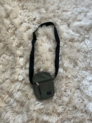 Grön axelväska med svart rem - Snygg liten axelväska i militärgrönt tyg med svart justerbar axelrem. Väskan har en dragkedja framtill och ett huvudfack med svart innerfoder och innerficka. Perfekt för att bära det viktigaste och enkel att matcha till olika outfits.💕💕💕