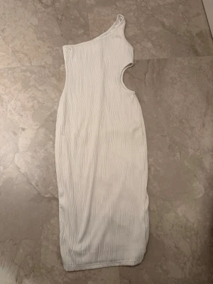 Vit one shoulder klänning - Trendig vit klänning med one shoulder-design och ribbad struktur. Klänningen har en snygg cut-out på sidan och är figurnära med en längre passform. Perfekt för dig som vill sticka ut med en modern och stilren look.