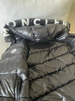 Svart väst från Moncler - Snygg svart väst från Moncler med glansig finish och stor huva med logotyptext. Jackan har dragkedja, klassisk quiltad design och ikonisk Moncler-patch på ärmen. Insidan har ett serietryck och kvalitetsmärkning. Perfekt för kalla vinterdagar.