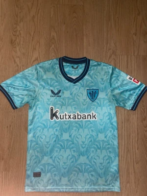 Ljusblå Athletic Club Bilbao tröja - Snygg ljusblå matchtröja från Athletic Club Bilbao med mörkblå detaljer och mönster över hela tyget. Tröjan har klubbmärke på bröstet, sponsorlogga 'Kutxabank' och Castore-logga. Tillverkad i lätt och ventilerande material, perfekt för fotboll eller träning.