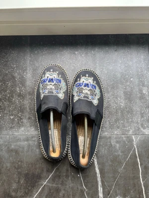 Svarta Kenzo loafers med tigerbrodyr - Säljer ett par svarta loafers från Kenzo med ikonisk tigerbrodyr i blått, vitt och beige framtill. Skorna har en klassisk slip-on design och kontrastsömmar runt sulan. Tillverkade i canvas med detaljerad brodyr som ger en unik och trendig look. Pris kan diskuteras vid snabb affär 