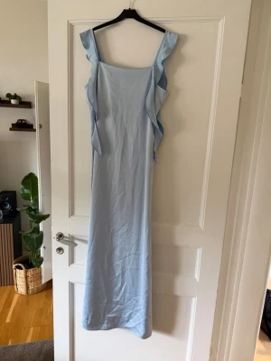 Ljusblå långklänning med volang - Elegant ljusblå långklänning med breda axelband och volangdetaljer längs sidorna. Klänningen har en rak siluett och är ärmlös, vilket ger en stilren och modern look. Perfekt för sommarens speciella tillfällen.
