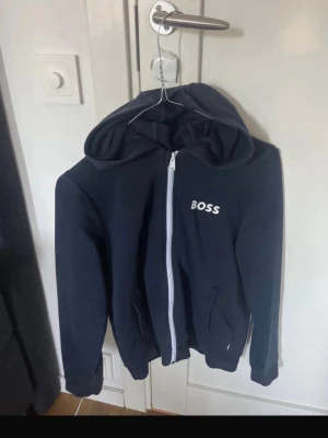 Svart hoodie med dragkedja från BOSS - Snygg svart hoodie från BOSS med vit dragkedja och logga på bröstet. Hoodien har huva, två fickor framtill och ribbade muddar vid ärmslut och nederkant. Perfekt för en avslappnad och stilren look. Det är främst mot pojkstorlek! Upp till 160cm 
