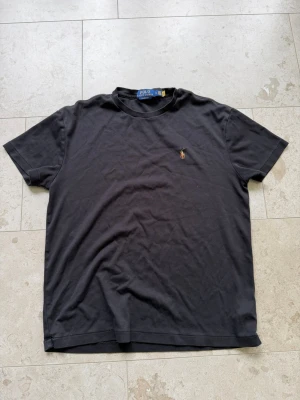 Svart t-shirt från Polo Ralph Lauren - Klassisk svart t-shirt från Polo Ralph Lauren med rund hals och broderad logga på bröstet. Modellen är enkel och stilren, perfekt till jeans eller shorts. Tillverkad i mjuk bomull för skön känsla hela dagen.
