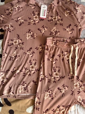 Blommig rosa/peach pyjama - Mysig pyjamas i två delar. Setet är ljusrosa med blommigt mönster. Tröjan har korta ärmar och krage, byxorna har resår i midjan och dragsko. Materialet är ribbat och mjukt för extra komfort. Jättefin pyjama och helt ny!!