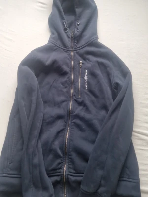Marinblå hoodie från Sail Racing - Snygg marinblå hoodie från Sail Racing med dragkedja framtill och en bröstficka med logga. Hoodien har justerbar huva och är tillverkad i mjukt, fleece-liknande material som känns skönt mot huden. Perfekt för en avslappnad och sportig stil.