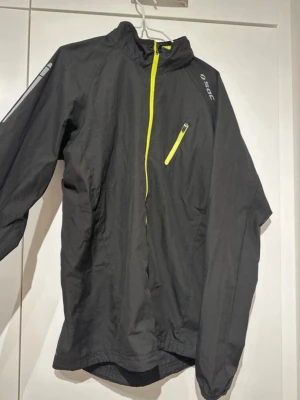 Svart vindjacka från SOC med gul dragkedja - Snygg svart vindjacka från SOC med gul dragkedja och bröstficka. Jackan har hög krage, långa ärmar och en sportig look. Perfekt för blåsiga dagar och enkel att packa ner i väskan. Reflekterande detaljer på axeln för extra synlighet.