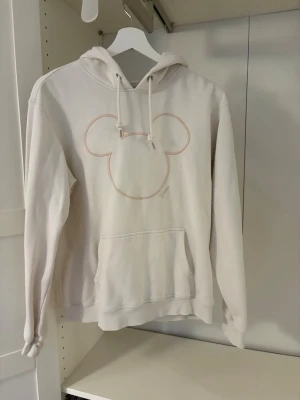Vit hoodie med Mickey Mouse-tryck - Säljer en vit hoodie med Mickey Mouse-tryck i ljusrosa på bröstet. Hoodien har huva med dragsko och en magficka. Från HM. 
