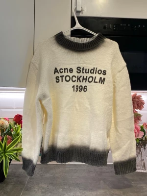 Vit stickad tröja Acne Studios - Stickad tröja från Acne Studios i vitt med grå ribbstickade muddar, nederkant och krage. Texten 'Acne Studios STOCKHOLM 1996' är tryckt framtill. Oversized passform och mjukt material, perfekt för en trendig och avslappnad look.