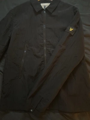 Svart overshirt från Lyle & Scott - Stilren svart overshirt från Lyle & Scott med dragkedja framtill och två snedställda bröstfickor med dragkedja. Klassisk krage och ikonisk gul logga på ärmen. Tillverkad i ett lätt, strukturerat material som ger en modern look.