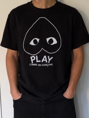 Svart Comme des Garçons Play t-shirt - Säljer en svart t-shirt från Comme des Garçons Play med det ikoniska hjärtat med ögon tryckt i vitt på bröstet. T-shirten har rund hals och korta ärmar. Perfekt för dig som gillar streetwear och vill ha en stilren men ändå unik look.