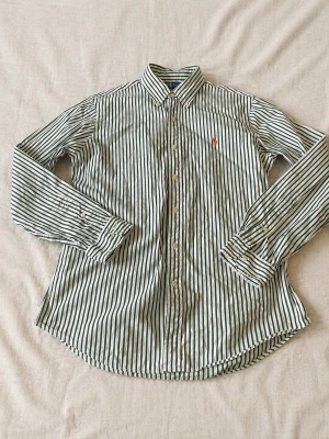 Randig skjorta från Ralph Lauren - Klassisk vit skjorta med gröna ränder från Ralph Lauren. Custom fit-modell med lång ärm, knappar framtill och ikonisk broderad logga på bröstet. Perfekt för dig som gillar stilrena och tidlösa plagg.