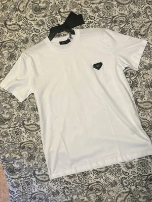 Vit t-shirt från Prada - Snygg vit t-shirt från Prada med den ikoniska svarta trekantsloggan på bröstet. Klassisk passform och rund halsringning. Tillverkad i mjuk bomull som känns skön mot huden. Perfekt för dig som gillar stilrena och exklusiva plagg.