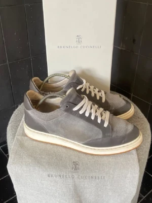 Grå sneakers från Brunello Cucinelli - Snygga grå sneakers från Brunello Cucinelli med ovandel i mocka och läder, vita snören och en vit sula med beige detalj. Klassisk låg modell med perforerade detaljer på tån och stilren design. Perfekta för dig som gillar exklusiva och tidlösa sneakers.