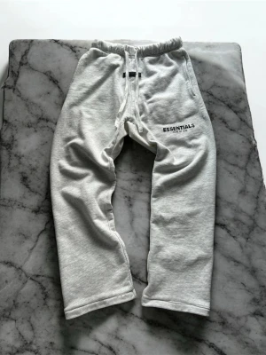 Gråa Essentials mjukisbyxor - Snygga grå mjukisbyxor från Fear of God Essentials med svart logga på vänster lår. Byxorna har resår i midjan med dragsko och raka ben. Perfekta för en chill och avslappnad stil. Tillverkade i mjukt material för maximal komfort. Uncuffed, skick 8/10 finns märke på snöret som såkert går bort i tvätten.