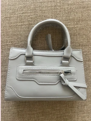 Ljusgrå minihandväska med dragkedja - 💓Trendig ljusgrå minihandväska i slätt konstläder med silverfärgad dragkedja framtill och dekorativa sömmar. Väskan har både handtag och en avtagbar axelrem för olika stilar. Perfekt för dig som gillar minimalistisk design och vill ha en liten men snygg accessoar.💗