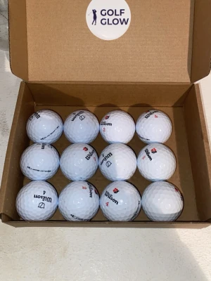 Wilson Duosoft 12-pack  - Säljer ett set med tolv vita golfbollar från Wilson, levererade i en brun kartong med etiketten 'Golf Glow' på insidan av locket. Bollarna har klassisk dimple-yta och märkning i svart och rött.