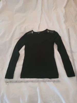 Svart långärmad topp med dragkedjor - Ralph Lauren, Stilren svart långärmad topp med coola dragkedjedetaljer vid axlarna. Tight passform och rund halsringning. Perfekt att matcha med jeans eller kjol för en trendig look.