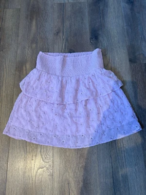 Ljuslila volangkjol med broderi - Söt ljuslila kortkjol med dubbla volanger och broderade blommor. Kjolen har bred resår i midjan för skön passform och är perfekt för vår och sommar. Tillverkad i mjuk bomull som känns lätt och luftig.