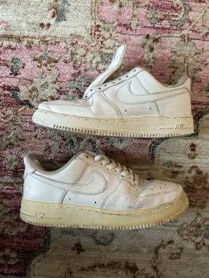 Vita Nike Air Force 1 sneakers - Klassiska vita Nike Air Force 1 sneakers i läder med låg profil och ikonisk swoosh på sidan. Skorna har en tjock, beige sula och perforerade detaljer på tån för extra ventilation. Perfekta för dig som gillar streetwear och tidlös stil.