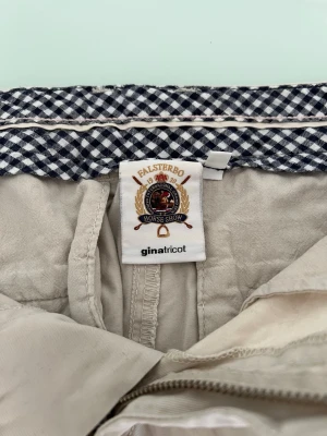 Beige shorts från Gina Tricot - Snygga beige shorts från Gina Tricot med vikta benslut och bälteshällor. Insidan har ett rutigt mönster i midjan för en extra detalj. Perfekta för varma dagar och enkla att matcha med olika toppar.