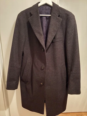 Grå rutig rock från Turo - Klassisk grå rock från Turo med diskret rutmönster. Rocken har tre knappar framtill, breda slag och två snedställda fickor. Fodrad insida med innerficka och Turo-logga. Perfekt för en stilren look under kallare dagar.