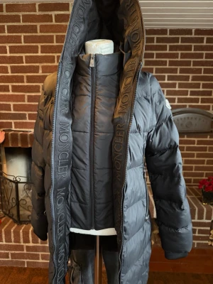 Svart Moncler dunjacka  - Snygg svart dunjacka från Moncler med stor, vadderad huva och dragkedja framtill. Jackan har puffig design, långa ärmar och Moncler-logga på huvan och ärmen. Perfekt för kalla vinterdagar och riktigt trendig look.